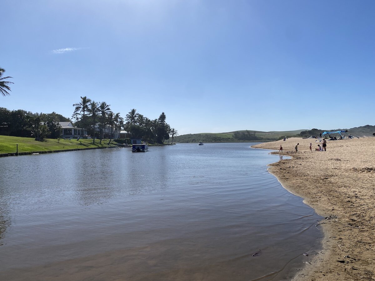 People enjoying the lagoon and sandy beach in Zinkwazi, with palm trees and distant hills under a clear sky.