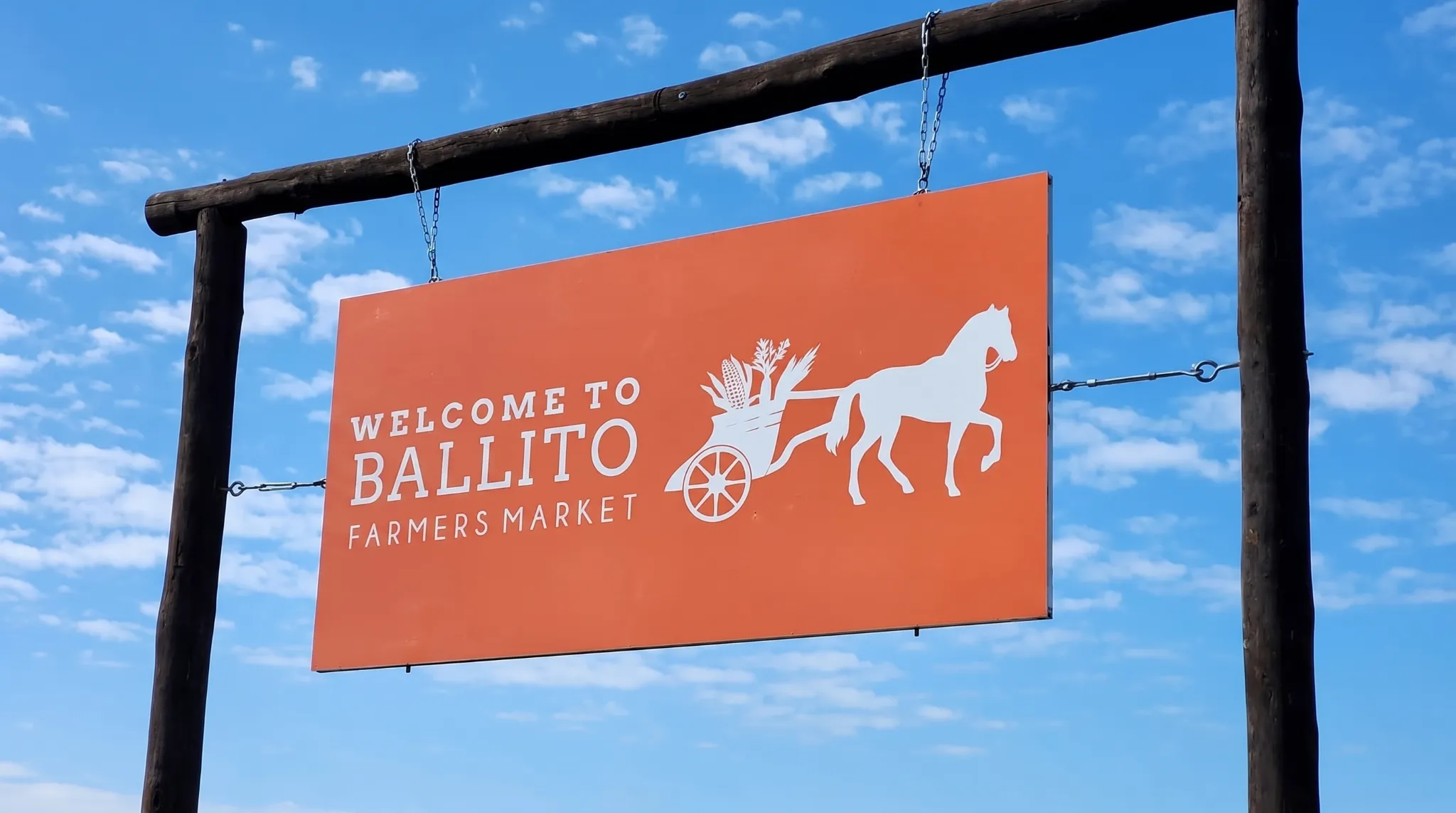 An orange sign welcomes visitors to the Ballito Farmers Market, featuring a horse pulling produce under a blue sky.