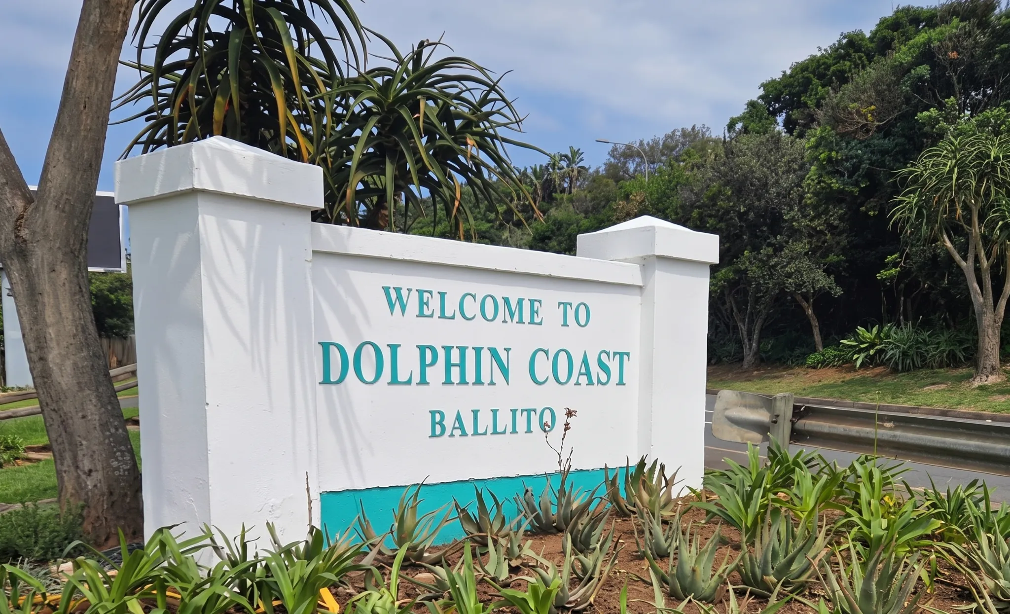 A white welcome sign for Dolphin Coast Ballito, surrounded by lush green foliage and trees.
