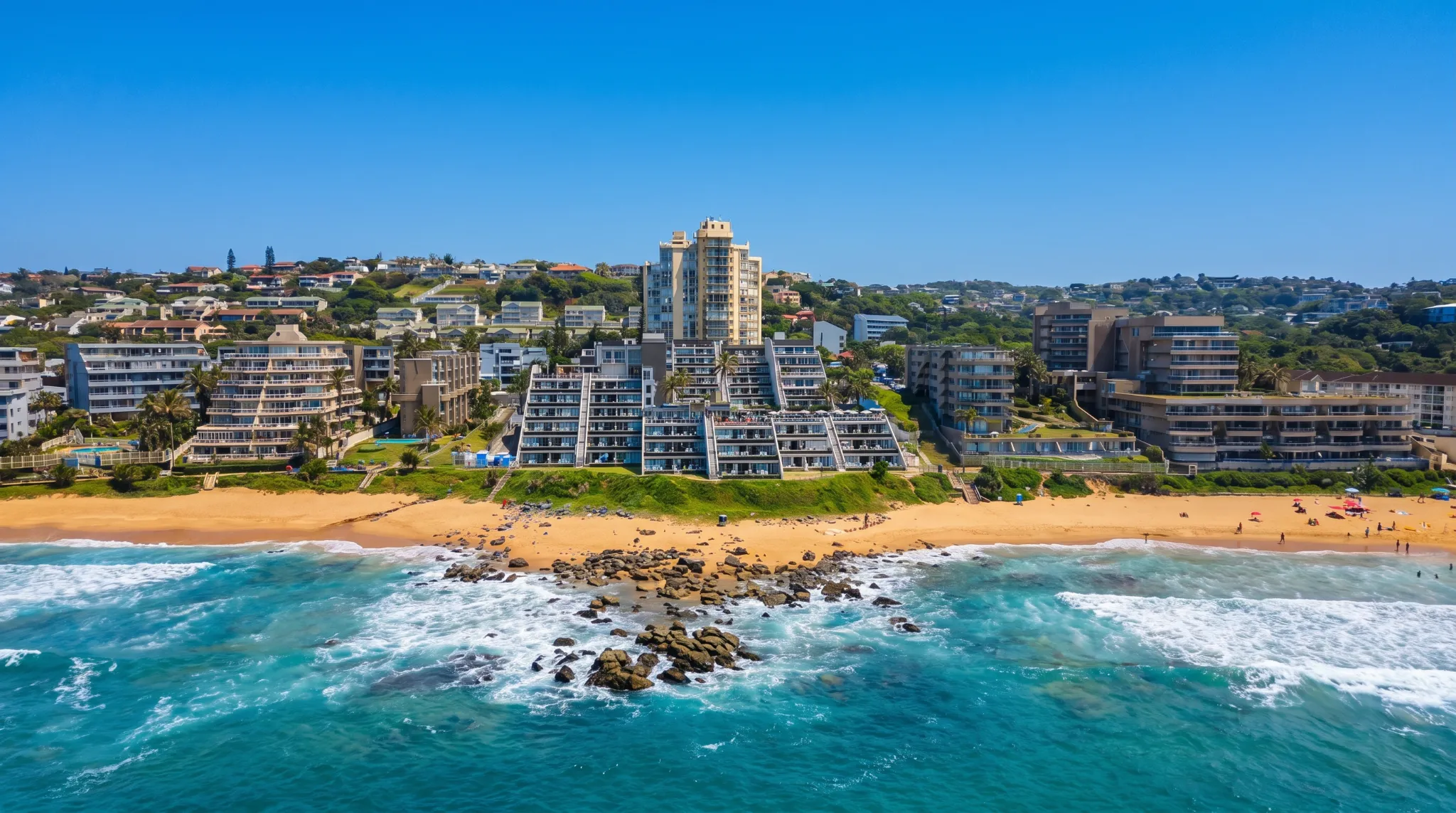Aerial view of Ballito's coastline, with buildings rising behind a sandy beach, rocks, and blue ocean waves.