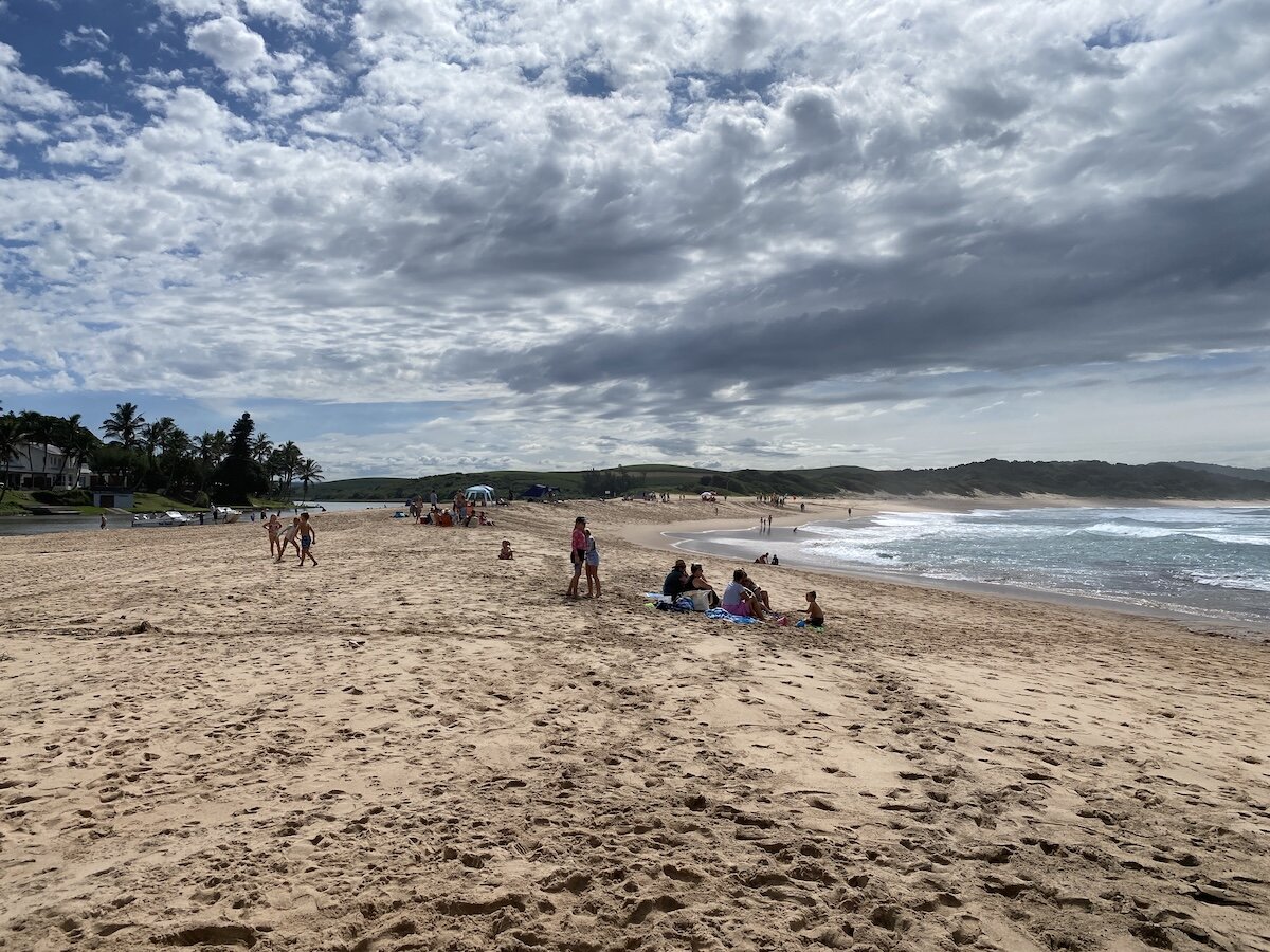 Families relax on the expansive sandy beach with the ocean and lagoon under a cloudy sky at Zinkwazi.