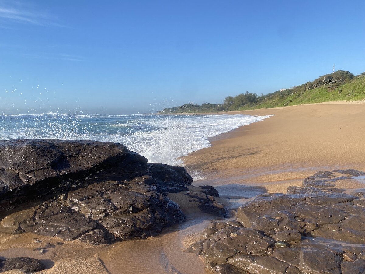 Waves crash on dark rocks and golden sand at Zinkwazi Beach under a clear blue sky.