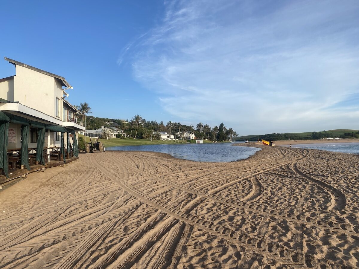 Tire tracks on the sandy beach leading to the lagoon in Zinkwazi with buildings and palm trees nearby.