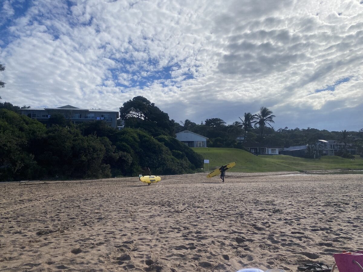 People with a surfboard and beach toys on a sandy beach at Zinkwazi with houses nestled in lush greenery.