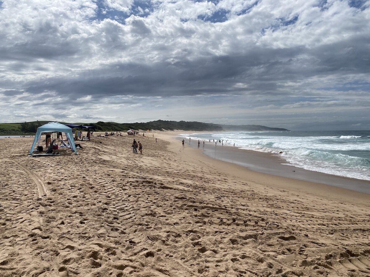 Cloudy day at Zinkwazi beach with people enjoying the ocean and sandy shore under a blue striped canopy.