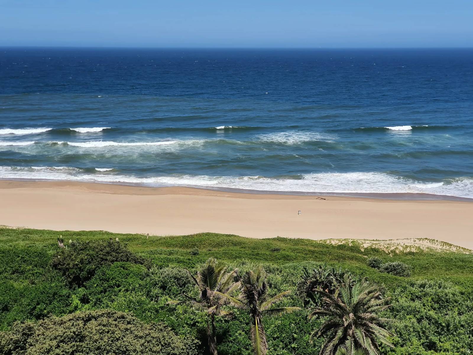 Ocean waves hit a pristine sandy beach framed by lush green vegetation and palm trees in Ballito.