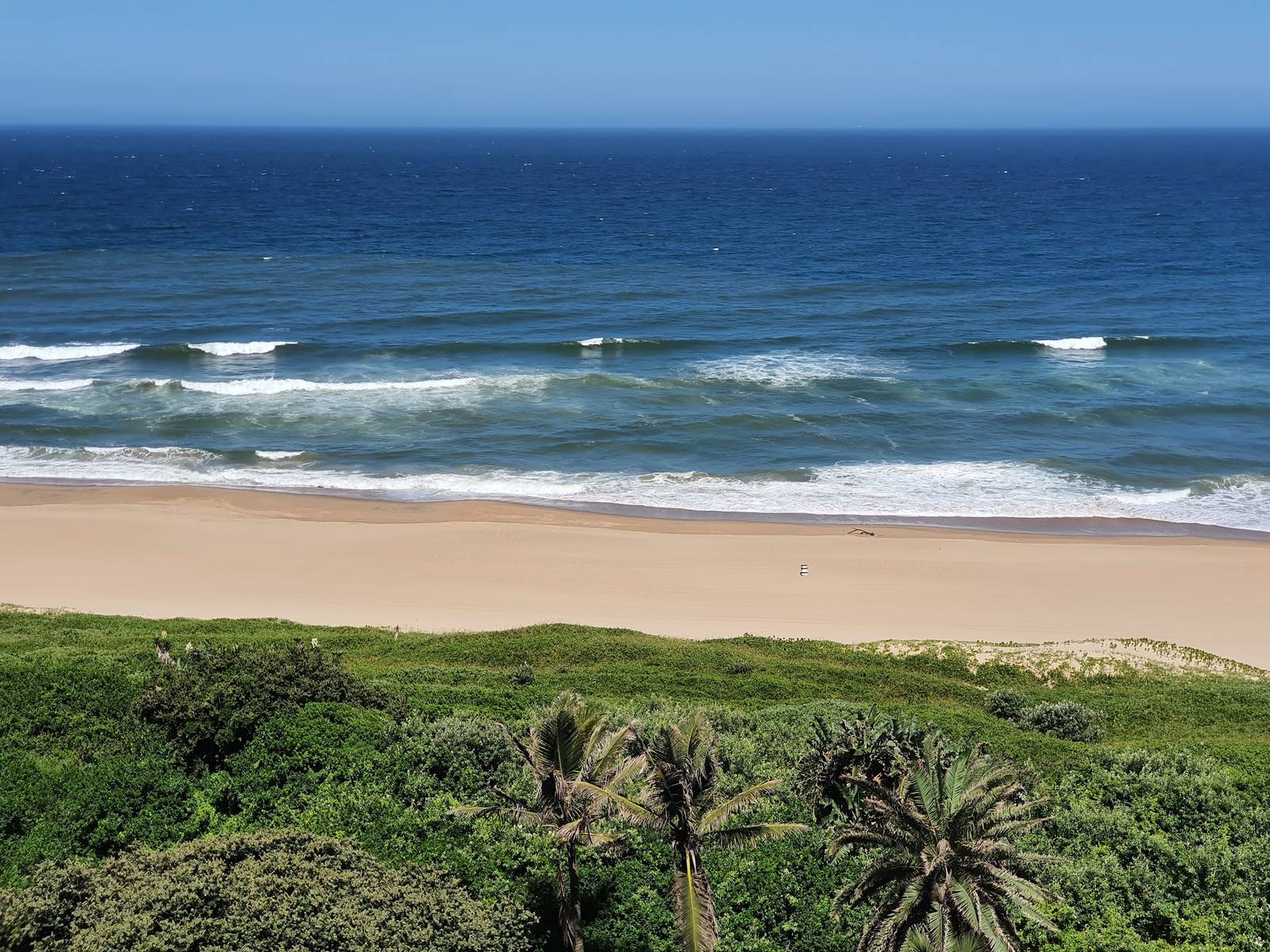 Ocean waves hit a pristine sandy beach framed by lush green vegetation and palm trees in Ballito.