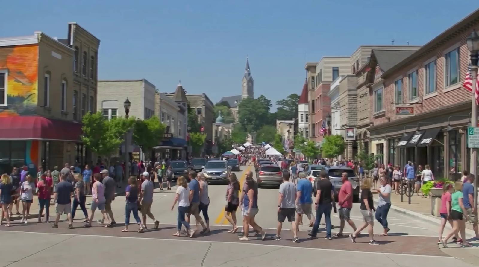 Crowds of people cross a street lined with historic buildings and cars on a sunny day in Port Washington.