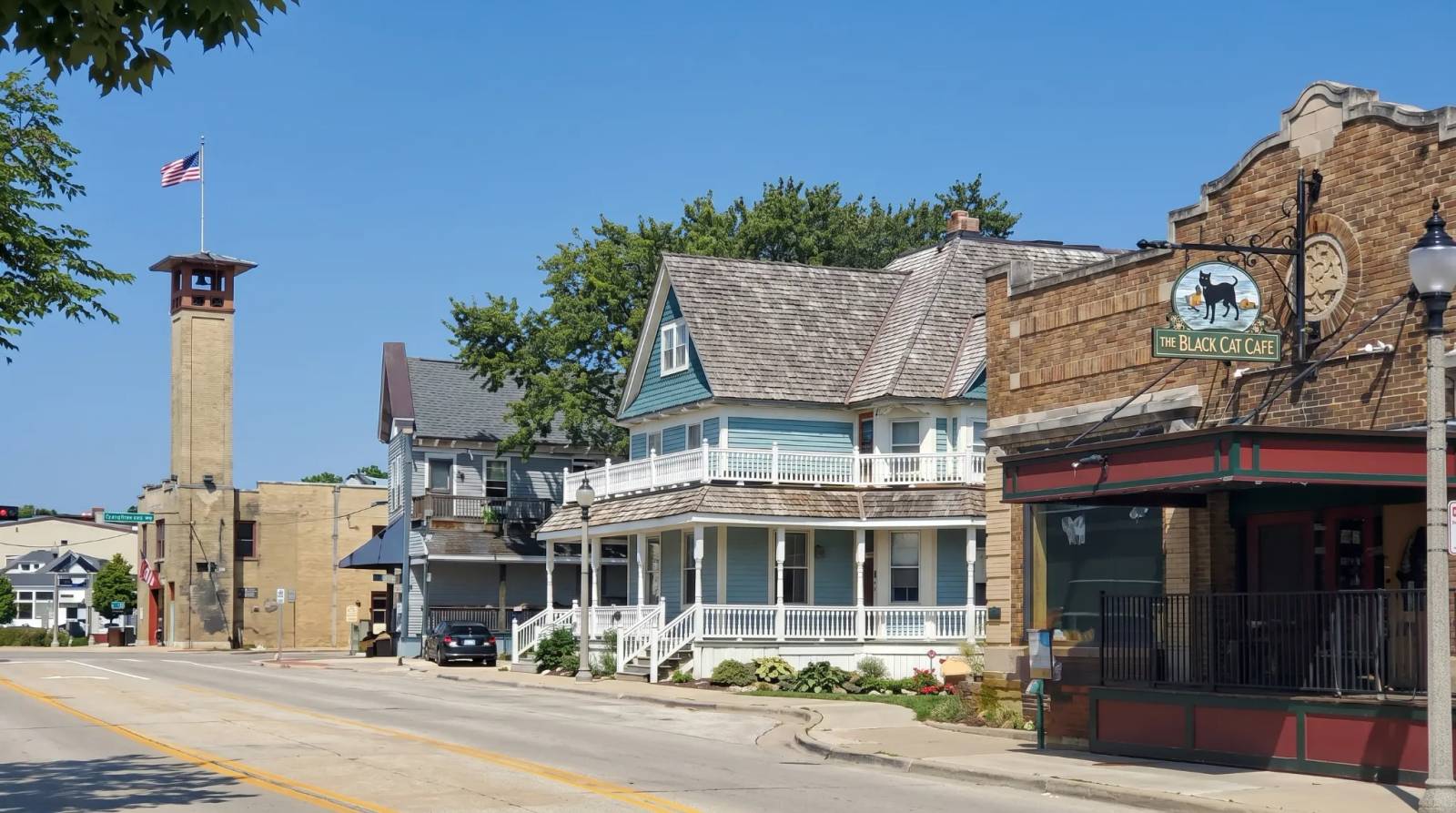 Main Street in Thiensville, featuring a building with intricate porch railings, a brick tower, and green trees.