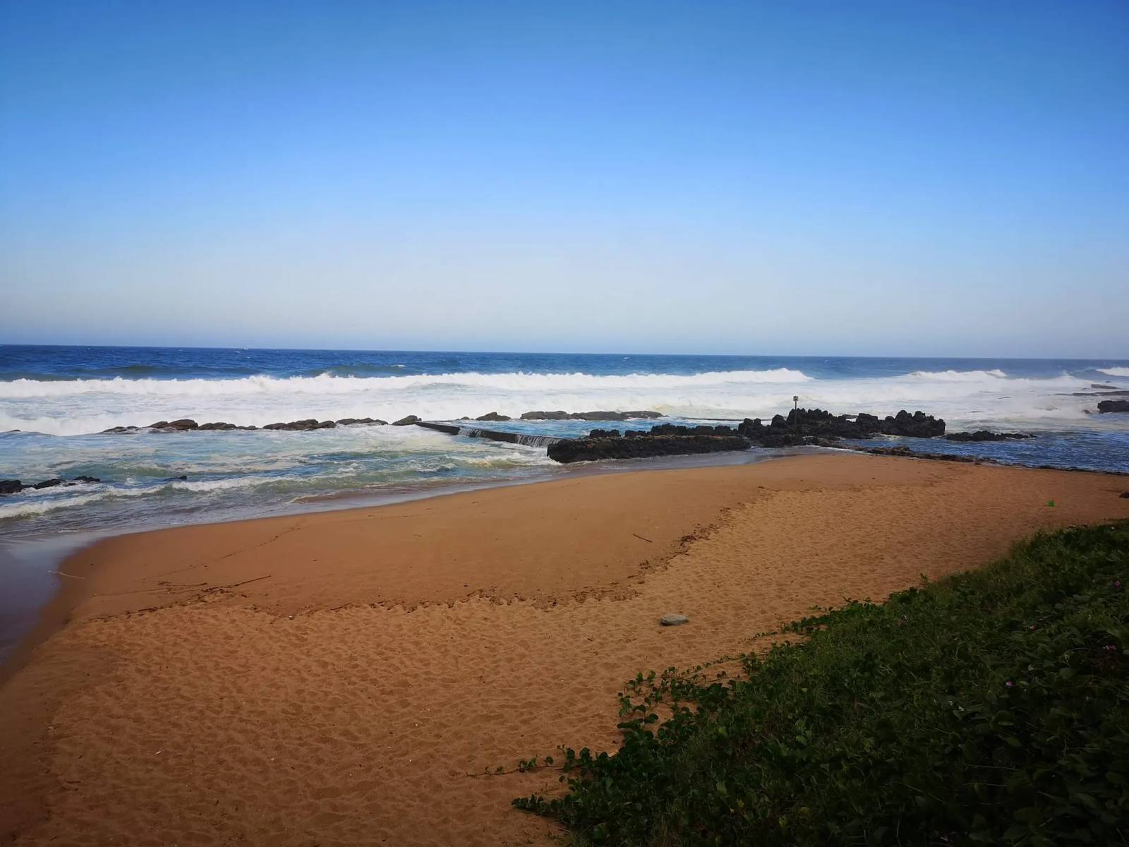 Waves breaking over a sandy beach and tidal pool at Shaka's Rock under a clear blue sky.