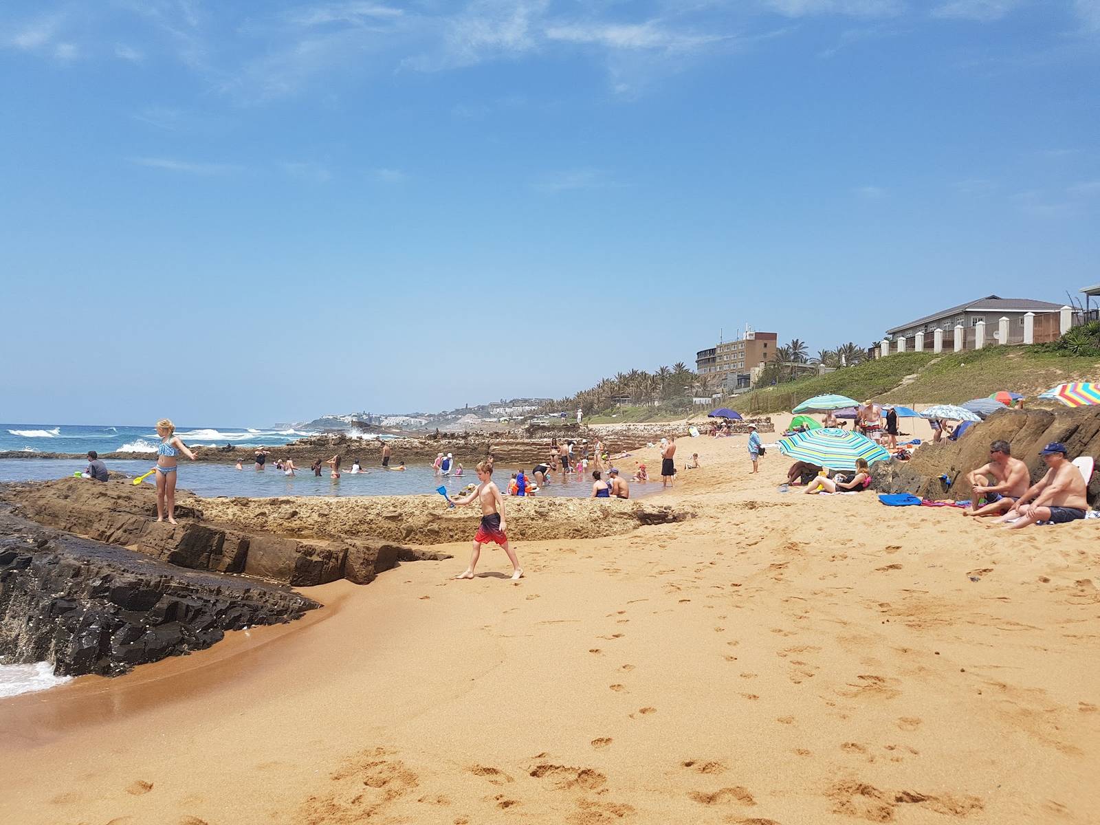 A sandy beach with people swimming in a rock pool and walking along the shore at Salt Rock.